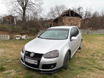 A silver Volkswagen Golf Mk5 GTI, bearing Bulgarian license plate EB 8303 BM, is positioned on a patch of uneven, sparse grass in Gorna Rositsa, Bulgaria. The vehicle's front and left side are prominently featured, with its metallic surface appearing wet, indicative of recent precipitation. Black rain visors are affixed to the windows, and a black lower front spoiler is visible. The immediate background, under a uniformly overcast sky, reveals a semi-rural environment. To the left, stacks of grey concrete blocks are present on an embankment. A large, bare deciduous tree stands centrally in the mid-ground, partially obscuring a distant residential building. Directly behind the vehicle, a derelict single-story brick structure with a severely damaged or collapsed roof is visible. To the right of this structure, a partially constructed concrete block wall extends horizontally. A faint human figure is discernible standing amongst leafless trees in the background, near the derelict building. The overall vegetation consists of sparse, bare trees and bushes on elevated, uneven terrain.