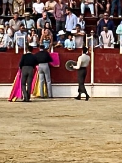 The image shows a bullfighting scene in what appears to be a traditional bullring. Three bullfighters, dressed in traditional attire, are visible. Two of them stand together near the barrier holding capes, while the third is further away and carries what appears to be a round metal object, likely part of the equipment used in the bullfight. A crowd of spectators fills the stands in the background. Based on the setting and the activity, this is likely taking place in the city of San José del Valle, Spain.