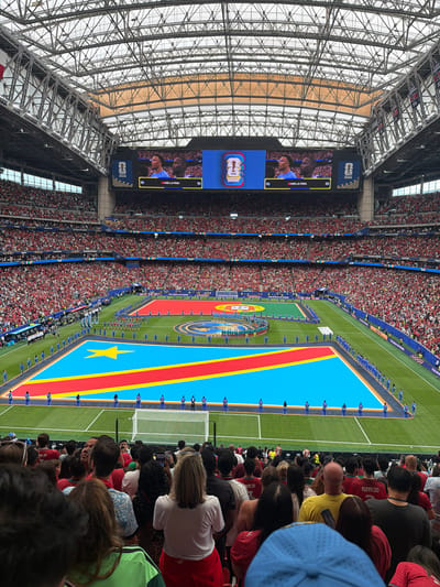 This media file captures a scene inside a large, modern stadium, likely during a sporting event. The primary subjects are a crowd of people filling the stands and individuals on the field. The field itself is a vibrant green football pitch marked with white lines. Dominating a significant portion of the field is a large flag, appearing to be the flag of the Democratic Republic of Congo, with its distinct blue, red, and yellow colors and a yellow star.
On the field, rows of people, likely players or officials, stand in formation along the edge of the flag. They are dressed in matching blue uniforms. In the background, the stadium's massive, intricate steel roof structure is visible, with a clear, possibly retractable, roof allowing natural light to enter. Large screens are mounted within the stadium, displaying graphics and images. One screen shows two individuals, possibly athletes, with the text "DANS LA PAIX" (meaning "In Peace" in French) displayed. Another screen prominently features the "FIFA WORLD CUP 2026" logo.
The crowd in the stands is densely packed, with many people wearing red, suggesting a possible team affiliation or theme for the event. Some individuals in the crowd are wearing jerseys with names like "RONALDO" visible. The overall atmosphere appears to be one of anticipation and excitement, common at major sporting events. The lighting suggests it is daytime. The presence of the FIFA World Cup 2026 branding and the context of Houston, USA, indicate this is likely a preparatory event or part of the build-up to the tournament.