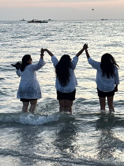 The image depicts three individuals standing in the shallow waters of Cenang Beach, Malaysia, at twilight. All subjects are partially silhouetted. The leftmost individual wears a white tunic with dark floral patterns and dark shorts, holding a dark hat in their left hand, and is partially turned to their right with a visible facial profile. The central and rightmost individuals wear white tops and dark shorts, facing away from the viewer. All three have their arms raised overhead, holding hands with each other, forming a raised arc. Water levels around the subjects range from knee-high to mid-thigh, with small waves visible breaking around their legs. The ocean extends into the background under a pale sky. Multiple distant watercraft are present, including a seaplane or similar aircraft on the water to the left, and other vessels further out. A single parasail is visible in the sky towards the upper right.