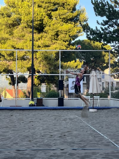 A male beach volleyball player is captured mid-air in a dynamic pose, with his body arched backwards and arms extended, as if preparing to hit or having just hit a ball. He is wearing a purple and black tank top and black shorts. The action is taking place on a sandy beach volleyball court, with a white net in the background. Behind the net, there are tall green trees, a white fence, and buildings that suggest an urban or semi-urban setting. Two white chairs and a closed beach umbrella are visible near the net. The sky is blue and clear. The location is specified as Gošići, Montenegro.
