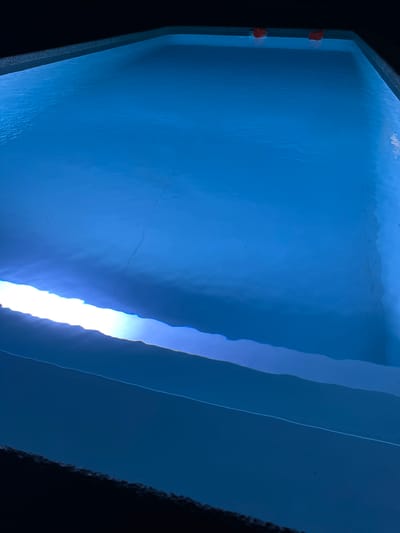 The image shows an illuminated swimming pool in the city of Izobilnyy, Russia. The pool is filled with blue water, and a bright light shines across the surface, creating a luminous band. The edges of the pool are visible, and in the background, two orange objects are partially submerged. The overall scene suggests a tranquil and perhaps private setting, illuminated for nighttime use.
