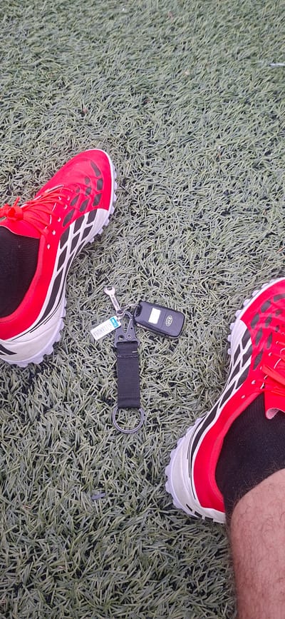 The image presents a top-down view of a person's feet and car keys resting on an artificial turf surface. The subject is a person, with portions of their lower legs and feet visible. They are wearing black socks and striking red and white athletic shoes, consistent with turf shoes or soccer cleats, suitable for artificial grass. The right leg shows some hair.

Positioned between the feet on the turf is a black Kia car key fob. It is attached to a keychain ensemble that includes a black fabric strap, a metal carabiner-style clip, and a large metal split ring. Also connected to the keychain is a small, silver-colored metal charm shaped like a wrench, bearing the inscription "DIAMOND®". A small blue and white tag is also part of the keychain, with the word "LUCKY" clearly visible at the top, followed by an unreadable alphanumeric string.

The setting is an outdoor artificial turf sports field or pitch, characterized by its uniform green synthetic grass blades. The lighting suggests it is daytime, casting a bright illumination without strong, defined shadows. The presence of athletic footwear on a sports field implies the person is engaged in, or pausing from, a sporting activity. The scene is set in Lyaskovets, Bulgaria.