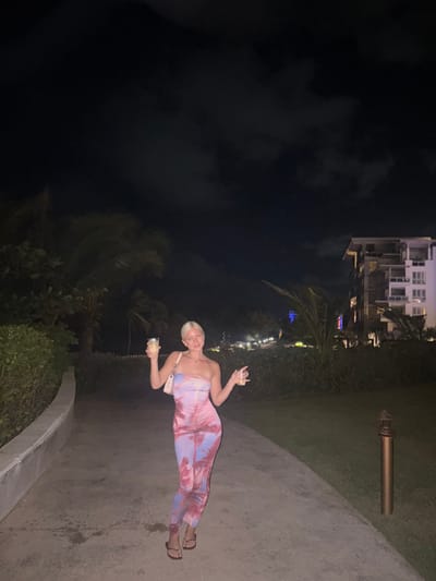 A woman poses on a walkway at night, holding a drink in each hand. She wears a one-shoulder, pink-and-blue patterned jumpsuit and sandals. A small, white shoulder bag is slung across her body. In the background, there is a building and trees. A dark sky fills the upper left portion of the image. A pipe is visible along the walkway, near a grassy area.