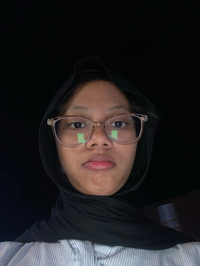 A close-up, low-angle selfie of a young woman wearing a black hijab and glasses is captured against a dark, out-of-focus background. The woman's face is illuminated from the front, highlighting her features and the light reflections on her glasses. She is wearing a light blue and white striped collared shirt. The image appears to have been taken indoors, possibly at night, with no specific identifiable landmarks of Tanggul, Indonesia visible.