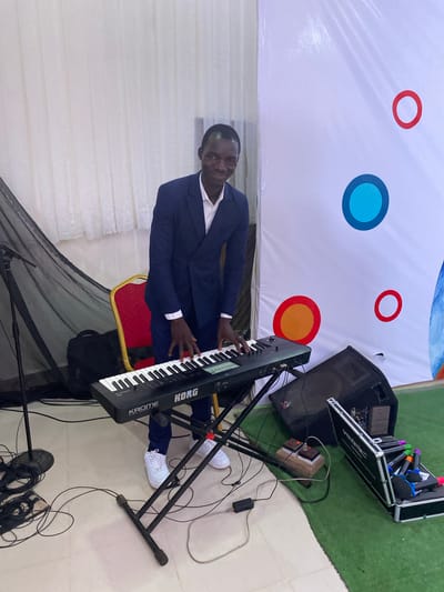A man in a dark blue suit, white shirt, and white sneakers stands behind a Korg keyboard. He is positioned in front of a white backdrop adorned with colorful circular designs. To his right, stage equipment includes a speaker, a mixer in a carrying case, and several microphones in a separate container. The floor is light-colored, and there is green Astroturf in the background. This scene is likely in Miango, Nigeria.