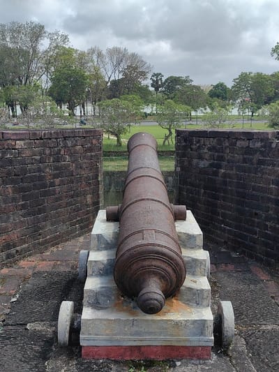 A rusty, dark brown cannon is positioned centrally, pointing forward, resting on a multi-tiered grey stone carriage with two visible grey wheels. It is flanked by two dark red-brown brick walls, which show signs of age. The immediate ground is paved with dark, weathered stones, and a red base is visible beneath the cannon's carriage. Beyond the brick walls, a narrow grassy area and a water-filled moat or trench separate the foreground from a paved road. The road features white markings and a parallel white barrier. In the mid-distance on the road, small figures on what appear to be motorbikes or bicycles are discernible, including a distinct dark blue-green scooter. The background is dominated by a dense line of trees with varying foliage, some green and others bare, with a large body of water visible behind them. The sky is uniformly overcast and grey. This scene is located in Phú Xuân Ward, Vietnam.