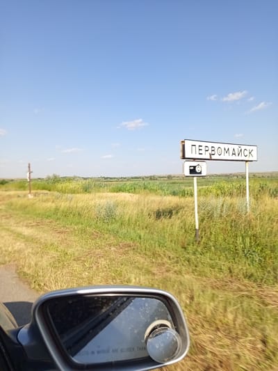 A roadside view is captured with a clear blue sky overhead, scattered with a few white clouds. In the middle ground, a field of tall green and yellow grass stretches towards the horizon. A wooden cross with a white figure is visible to the left. A signpost in the center right of the image bears the Cyrillic text "ПЕРВОМАЙСК," indicating the city of Pervomaisk, Moldova. Below this sign is a smaller sign with a camera icon. In the foreground, a portion of a car's side mirror is visible, reflecting the sky and a small convex mirror. The overall scene depicts a rural roadside environment on a sunny day.