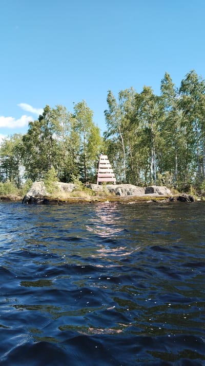 A bright, sunny day with a clear blue sky and scattered clouds is depicted. The scene is a body of water, likely a lake or river, with dark blue, choppy water in the foreground. In the background, there is a rocky island covered with green grass and a dense line of tall, green trees, primarily birch trees with white trunks. A distinctive red and white tiered structure, resembling a large ladder or a navigational marker, stands on the rocks in the middle ground, positioned among the trees. The reflection of the structure and the trees can be seen on the surface of the water. There are no people visible in the image. The overall impression is a natural, serene landscape.