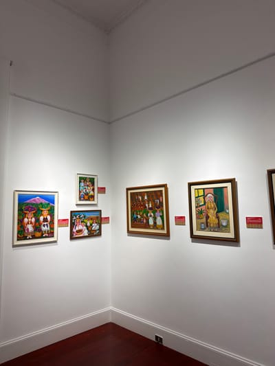 This is a photograph of an art exhibition in La Orotava, Spain, showcasing several paintings displayed on a white wall. The paintings appear to be folk art or naive art, characterized by vibrant colors and depictions of people engaged in daily activities, often with a rural or harvest theme.

On the left wall, there are three framed paintings. The largest one at the bottom left features two women with baskets of fruit on their heads, with a volcano in the background. Above this, a smaller painting shows a group of people carrying baskets. To the right of these, a rectangular painting depicts children and adults with baskets of fruit.

On the adjacent wall, there are two more framed paintings. The one on the left shows a group of people harvesting from trees. The painting on the right is a portrait of an older woman sitting in a chair, wearing a yellow dress and holding a basket. Red informational plaques are visible below some of the paintings.

The exhibition space has white walls and a dark wooden floor. The corner of the room is visible, and there is subtle architectural molding along the ceiling. The lighting appears to be artificial, illuminating the artwork. The overall impression is that of a well-maintained gallery space.