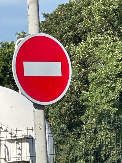 The media file shows a close-up of a red circular sign with a white rectangle in the center. The sign appears to be a traffic sign, possibly indicating "Do Not Enter" or a similar restriction, common in Ukraine. The sign is mounted on a grey, textured pole or barrier. In the background, there is lush green foliage from trees or bushes, and a black metal fence. The sky is visible in the upper left portion of the image, appearing blue. The lighting suggests it is daytime, with sunlight illuminating the scene and creating some highlights on the sign and foliage. The image is oriented vertically. No people are visible. The location context provided is Shatava, Ukraine.
