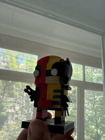 A hand holds a Lego creation in front of a window with sheer white curtains. The Lego object appears to be a stylized train engine, primarily red and yellow, with black accents and blue stripes. It has two white circular pieces that resemble eyes, and a black, curved piece on top that might be a hat or hair. A black, multi-pronged piece protrudes from the bottom of the Lego creation, resembling a mechanism or weapon. The background shows a window with several panes, through which dappled green foliage is visible, suggesting daylight and possibly a sunny day. The curtains diffuse the light, creating a soft, slightly hazy effect. The scene is indoors, with the natural light coming from outside. There is no visible text in the image. The overall mood is somewhat playful due to the Lego toy.
