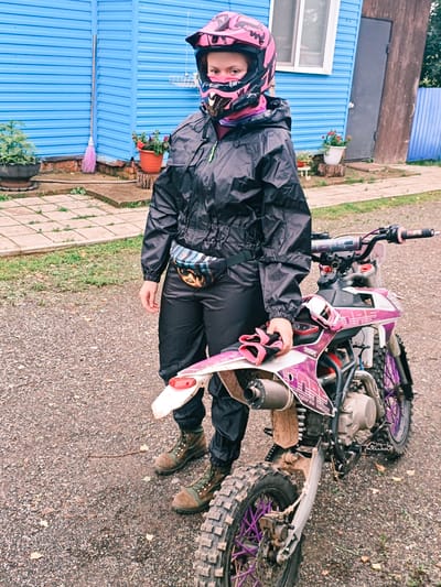 A young female with auburn hair is standing next to a dirt bike. She is wearing a black rain suit with a pink and black helmet. A fanny pack with a colorful pattern is around her waist. Her boots are brown and green. The dirt bike is predominantly pink and white with purple accents on the wheels. The background consists of a blue building, a fence, and some potted plants. The ground is covered in gravel. This scene is likely located in Rassol'naya, Russia, given the context provided.