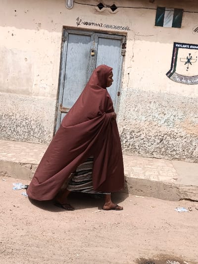An individual walks right-to-left across a dirt path in Zango, Nigeria, alongside a light-colored building wall. The person is dressed in a flowing, full-body maroon garment that covers their head and most of their form, revealing a horizontally striped dress or skirt underneath and dark flip-flops on their feet. The building wall features a light blue double door secured with a gold padlock. Above the door, black handwritten text reads "M.R- 08131991447, 08167685666." To the right of the door, a painted Nigerian flag is visible, alongside a crest featuring "VIGILANTE GROUP" and "Say No to Crim" text, with a crossed sword and rifle emblem. The ground is light brown dirt with scattered small debris. A raised concrete platform with a patterned surface runs along the base of the wall on the right side of the frame.