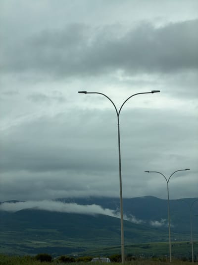The photograph, taken near Tedotsminda, Georgia, features a cloudy sky over a mountainous landscape. Dominating the foreground are a series of tall, grey streetlights with a distinctive "Y" shape at the top. The lighting fixtures are dark, contrasting with the overcast sky. In the background, a mountain range is partially obscured by low-hanging clouds, creating a layered effect. Green vegetation is visible on the lower slopes of the mountains, indicating a natural environment outside the immediate urban area.