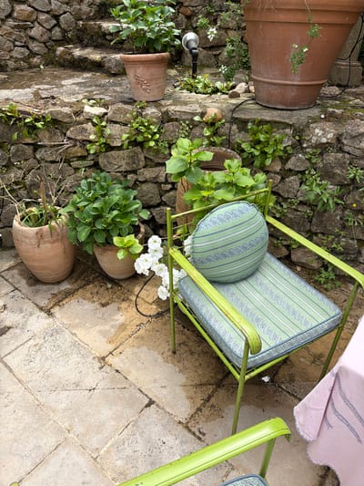 The scene is an outdoor setting in Saint-Victor-la-Coste, France. A light-green metal chair with a striped cushion and round striped pillow sits on a stone-tiled patio. The chair is positioned near a stone wall and several terracotta pots containing green plants. Some of the plants are flowering with white blossoms. A large terracotta pot is visible to the left of the stone wall, along with a small landscape lighting fixture. A portion of a pink tablecloth is visible in the upper right corner of the image.