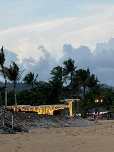 The image depicts a beach scene in Ban Bang Niang, Thailand. In the foreground, a sandy beach stretches towards the water, with faint footprints visible. Behind the beach, a rocky embankment supports two modern, illuminated structures with unique, branching designs. These structures are lit with warm yellow lights that create a striking visual against the dimming sky.
To the left of the illuminated structures, a metal staircase with railings leads up from the beach. Behind the structures, a dense line of tropical trees and palm trees forms a backdrop, with lush green foliage extending up a gentle slope. In the distance, a mountain range is faintly visible through the trees and clouds.
The sky is overcast with soft, diffused light, suggesting either early morning or late evening. A few distinct clouds are scattered across the sky. In the background, near the illuminated structures and trees, there are some lights visible, possibly from other buildings or streetlights. On the far right, a small section of what appears to be a building or structure can be seen.
There are a few scattered people visible in the scene. On the beach, near the right side, a person is sitting on the sand, and another person is standing further back. Near the illuminated structures, a red object, possibly a life preserver or buoy, is attached to one of the structures. A couple of kayaks or canoes are also visible near the edge of the beach on the right. The overall atmosphere is tranquil and tropical, with the artificial lighting of the structures adding a modern touch to the natural landscape.
