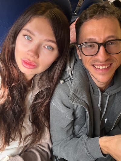 The image displays two individuals, an adult female and an adult male, seated in close proximity within what appears to be the cabin of a passenger aircraft, indicated by the blue upholstered seating visible in the background. The location is Colombier-Saugnieu, France.
The female, positioned on the left, has long, wavy dark brown hair, light blue-green eyes, and full lips, which are slightly parted. She is wearing a light beige or off-white ribbed top beneath a light brown or taupe zippered jacket, which features a circular dark button with a white 'B' emblem on the shoulder area. She looks directly towards the camera with a neutral expression.
The male, situated to the right and slightly behind the female, has short, dark, curly hair and is wearing dark-framed eyeglasses. He smiles broadly, revealing his teeth, and is dressed in a grey hooded sweatshirt with visible white stitching details on the seams. Both individuals are looking towards the camera. No specific actions beyond posing for the photograph are depicted.