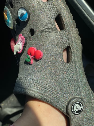 A close-up, eye-level shot shows a person's foot wearing a sparkly silver Croc shoe adorned with various charms. The charms include a cartoonish eye, a pair of cherries, and what appears to be a pink claw or hand. The Croc has the signature holes and branding. The background is dark, suggesting an indoor or shaded environment. The image does not provide any information about the city of Kfar Shmaryahu, Israel.