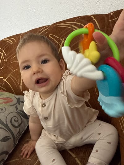 In this vertically oriented image, a baby is held and playing with a colorful toy. The baby, facing the camera, has bright blue eyes and an open mouth, suggesting excitement or surprise. They are wearing a light-colored, ruffled top and white leggings adorned with small pink hearts. The baby's left arm is extended, reaching for the toy, which is a multi-colored rattle with green, orange, yellow, red, and blue components.

The baby appears to be on a surface covered with a brown and beige patterned fabric, possibly a couch or blanket. A person's hand, with a pink fingernail, is visible at the top of the frame, holding the toy. The background includes a white textured wall on the left side and a patterned cushion with grey and floral designs on the lower right. The lighting suggests an indoor setting, likely during the daytime. The overall mood is playful and intimate, capturing a moment of interaction between the baby and the toy, with the implied presence of the person holding them. The image was taken in Momchilovtsi, Bulgaria.