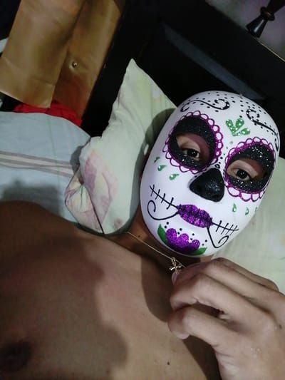 A person wearing a white sugar skull mask with glitter details and a gold necklace lies on a bed. The mask is decorated with purple and green accents. The person's eyes are visible through the mask's eyeholes. A hand is near the necklace. The background includes a bed with patterned sheets and a brown headboard.