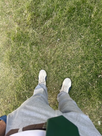 A first-person perspective shot shows a person standing on a patch of green grass. The person is wearing grey sweatpants and light-colored sneakers with dark blue accents. The grass is somewhat dry and sparse in places, with some lighter colored blades mixed with green. The scene appears to be an outdoor urban environment, likely a park or public space, possibly in the city of Kabal, Pakistan.