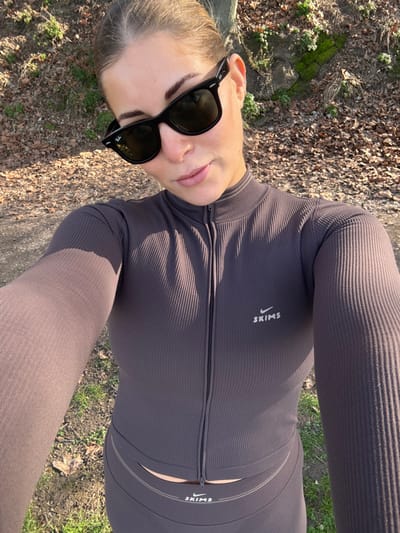The image captures a selfie taken outdoors by a woman on a bright, sunny day. The camera is held at a slightly elevated and angled position, looking down towards her upper body. The woman has fair skin and her hair, which appears light in color, is pulled back. She is wearing a pair of stylish, black rectangular sunglasses that cover her eyes, and a small silver earring is visible in her right ear. Her lips are a natural pink or nude shade, and her eyebrows are well-defined.

She is dressed in a cohesive dark brown or charcoal-colored ribbed athletic set. The top is long-sleeved, featuring a high mock-neck collar and a black half-zip closure down the front. Prominently displayed on the left side of her chest, facing the viewer, is the brand name "SKIMS" printed vertically in white capital letters, accompanied by a white stylized swoosh logo that resembles the Nike emblem. The top appears to be cropped, showing a slim section of her midriff above the waistband of her bottoms. She is wearing matching high-waisted leggings or pants, made of the same ribbed material and color. The waistband of the bottoms also features the white "SKIMS" text and swoosh logo on the left side, aligning with the top. Additionally, there are a couple of thin, light-colored horizontal lines integrated into the design of the waistband just above the "SKIMS" logo.

The background consists of an outdoor natural setting. The ground is covered with an assortment of dry brown leaves, dark soil, and scattered patches of green grass. The strong sunlight casts visible shadows across her clothing, indicating a clear sky and daytime conditions. The overall scene suggests a casual outing or active pursuit in comfortable, branded athletic wear.