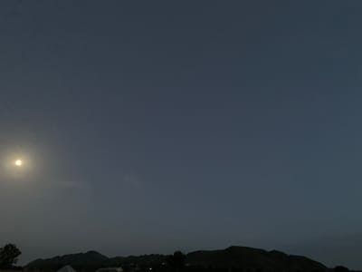 The image depicts a nighttime scene in Zawan, Nigeria. The sky is a deep, dark blue, with a bright, full moon shining prominently in the upper left quadrant. Below the moon, a faint, wispy cloud is visible. The lower portion of the image is dominated by the silhouette of a range of hills or mountains. The outlines of trees and structures are barely discernible against the dark landscape. The overall impression is of a tranquil, nocturnal environment.