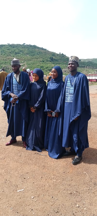 Four people are standing outdoors in Naraguta Hausawa, Nigeria. Two men and two women are dressed in blue, traditional Hausa clothing. The men wear embroidered caps and robes with patterned vests. The women wear hijabs and long dresses, also in blue. The ground is sandy. In the background, there is a hill covered in vegetation and a glimpse of a red and white structure.