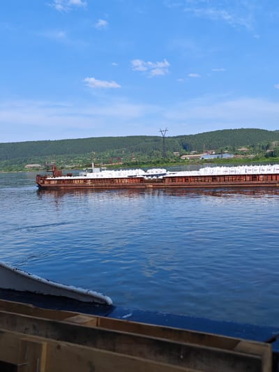 An outdoor, low-angle, medium shot captures a barge laden with white sacks on a wide, blue river. The barge, primarily a rusty red color with some white accents, is moving from left to right in the frame. The water's surface exhibits gentle ripples, reflecting the blue sky and scattered white clouds above. In the background, a lush green, tree-covered hill slopes down towards the river, dotted with small buildings and industrial structures, including a tall transmission tower. The foreground features the edge of another vessel, with visible wooden crates and a dark blue hull. The scene appears to be set in the city of Pristan', Russia, given the context of the barge traffic on the river and the surrounding landscape. There are no people visible in the image.