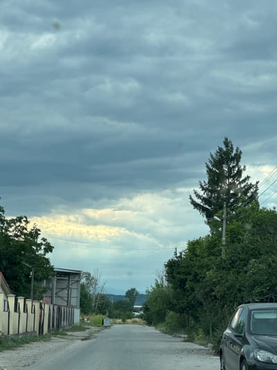 This is a vertical, medium-shot photograph taken from the perspective of someone in a car, looking out at a rural roadside scene. The sky dominates the left side of the frame, filled with dramatic, layered grey clouds interspersed with a bright, yellowish-white band of clouds, suggesting either sunrise or sunset with varying cloud cover.
On the right side of the image, a paved road runs diagonally into the distance. Along the side of the road, there is a dense line of green trees and vegetation. To the right of the road, a dark-colored car is partially visible in the upper right corner. Further down the road, there are several structures, including a building with a corrugated metal roof and a light-colored wall with barred windows, and a smaller grey bin. A utility pole with a light fixture stands near the trees. In the far distance, a hazy outline of mountains can be seen against the sky.
The overall impression is a slightly overcast day, possibly late afternoon or early morning, in a rural area. The sun's rays are breaking through the clouds in one section, creating a bright contrast. There is no visible text in the image. The scene is quiet and appears to be a view from a moving vehicle.