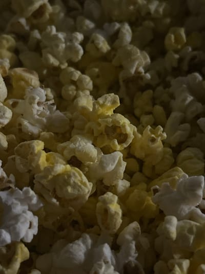 The image is a close-up, top-down view of a pile of popcorn. The popcorn pieces are varied in size and shape, with some appearing fluffy and white, while others are a pale yellow. The lighting is dim, creating shadows and highlighting the textures of the popcorn. There are no people, actions, or interactions visible in the image, and no specific location in Byala Slatina, Bulgaria can be determined from the provided visual information.