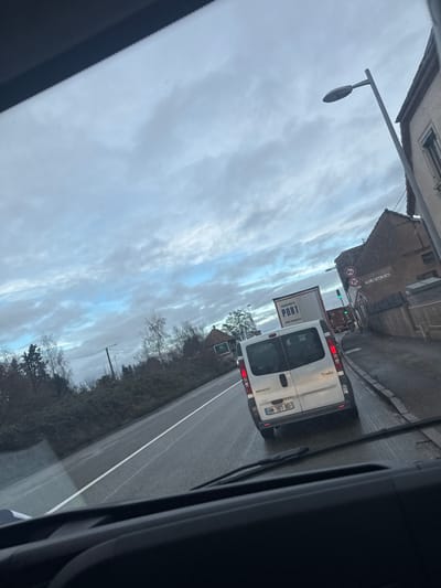 The image captures a road scene in Fegersheim, France, viewed through the windshield of a stationary or slow-moving vehicle. The sky is largely overcast with layered gray and white clouds, revealing some patches of lighter blue. The asphalt road surface is visibly wet. Directly ahead of the camera vehicle is a white Renault Trafic van, bearing French license plate BN-787-BD, with its rear exterior and unilluminated taillights visible. Further ahead, a white semi-trailer truck displaying "TRANSPORTS PORT" on its side is observed, followed by another truck, with a green traffic light visible in the distance.

To the right side of the road, a paved shoulder borders an older building with an orange-tiled roof and a light-colored facade, featuring a prominent window. Multiple road signs are installed along this side, including a "No Entry" sign (red circle with white bar), a "No Parking/Standing" sign (red circle with red diagonal bar), and a triangular warning sign with a pedestrian symbol. A light-gray streetlamp stands near the building. A partially visible sign on the building displays "TEL 0388". To the left of the road, a dense strip of winter vegetation consists of bare deciduous trees, green evergreens, and shrubs. A dark utility pole with overhead wires is also present on the left side of the road. No individuals are discernibly visible within the frame.