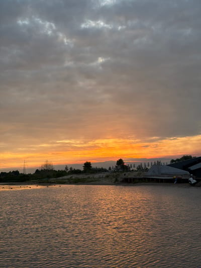 The image depicts a serene natural scene at dusk, likely at the "Sophiivka" beach and water park located in Khotianivka, Ukraine, as suggested by the context of previous images. The sky is dominated by a dramatic sunset, with vibrant shades of orange and yellow illuminating the horizon, transitioning to a muted grey and blue towards the top, where scattered clouds add texture. A body of water occupies the foreground, reflecting the warm hues of the sunset, with gentle ripples indicating a light breeze. Along the far bank, a sandy embankment is visible, lined with trees and some vegetation. A prominent electricity pylon stands on the left side of the landscape. Further to the right, structures resembling beach umbrellas are arrayed on a raised wooden platform, with a building partially covered by a grey tarp in the immediate vicinity. Several buoys are sparsely scattered in the water, marking a designated area. No people are present in the frame, and the overall atmosphere is tranquil and still.