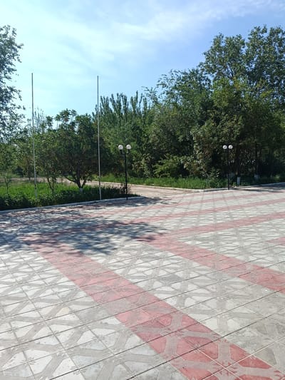 A paved plaza with red and white patterned tiles stretches towards a line of trees under a bright blue sky with sparse clouds. Two tall, slender poles stand vertically against the sky to the left of center, while two white globe-shaped lamps on a black pole are visible among the trees to the right. Shadows from unseen foliage dapple the patterned tiles. The location is identified as Kungrad, Uzbekistan.