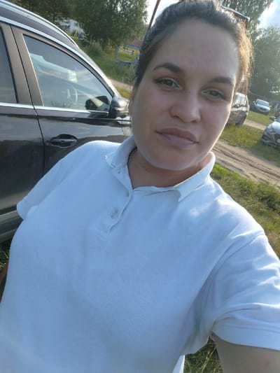 A middle-aged woman with dark hair pulled back is taking a selfie outdoors. She is wearing a white polo shirt with the collar turned up and a silver chain. She is standing next to a dark-colored car, and the background shows trees, a dirt road, and other vehicles. The image appears to have been taken in the city of Detskii Dom (Bashenino), Russia, on a sunny day.