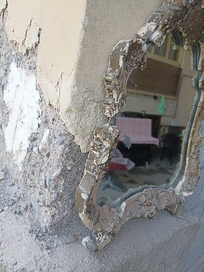 This is a close-up, angled shot of a heavily damaged, ornate mirror propped against a rough, crumbling concrete structure. The mirror's frame, once likely gilded, is chipped and peeling, revealing a textured, aged surface. The glass itself is also damaged, with a large crack running through the center and several dark smudges or accretions obscuring the reflection. In the reflection, a blurry interior scene is visible, featuring what appears to be a pink-covered bench or sofa and a green bag hanging on a wall. The concrete structure is textured with visible aggregate and areas where plaster or stucco has fallen away, exposing the rougher material beneath. The lighting suggests an outdoor, daytime setting. The location is stated to be Aygezard, Armenia.