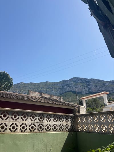 This is a photograph taken on a bright, sunny day in Montgó, Spain. The foreground features a green painted wall topped with a decorative concrete block fence with a repeating geometric pattern. Behind the fence, a low-lying building with a red facade and a tiled roof is visible. Further in the background, a white arched gateway with a tiled roof stands, leading to an area with trees and a black metal fence. Dominating the middle ground and stretching across the horizon is a large, rugged mountain range covered in sparse vegetation. The sky is a clear, bright blue with a few faint power lines stretching across it, and a thin contrail from an airplane is visible. A large, textured cactus is partially visible in the upper right and upper left corners of the frame, framing the scene. The lighting suggests it is mid-day, casting distinct shadows.