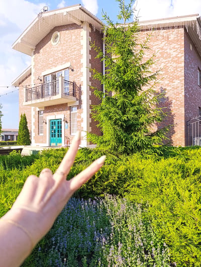 A medium shot shows a two-story brick house with a white trim roof and icicle lights hanging from the eaves. A large evergreen tree stands in front of the house, with a bed of bright green bushes and purple flowers in the foreground. In the lower corner of the frame, a hand is held up with two fingers extended in a peace sign, with a ring on one finger and a bracelet on the wrist. The scene appears to be outdoors, with a blue sky and white clouds visible in the background. The location is identified as Ivanovskoe, Russia.