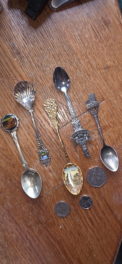 Six collectible spoons and three coins are arranged diagonally on a wooden surface. The spoons are varied in design. The first spoon from the left has a circular inset depicting a building and trees. The second spoon is shaped like a scallop shell. The third spoon is gold-colored and highly ornate. The fourth spoon is silver and features a sculpted building atop its handle, with text that appears to read "MUSEUM CATHEDRAL" and "DEUTSCHLAND". The fifth spoon is also silver and has a detailed, patterned handle. The sixth spoon is silver and has a plain, oval bowl. The coins are positioned to the right of the spoons. One coin is larger with a profile of a person and the number "50". Two smaller coins are also present. The wooden surface shows a prominent wood grain pattern. The scene is photographed from a top-down perspective. There is no indication of the city of Marum, Netherlands.