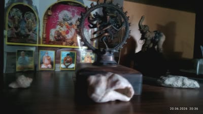 The image depicts an indoor setting with several religious and decorative objects arranged on a dark wooden surface. In the background, there are two framed pictures with figures of deities and what appear to be spiritual leaders or monks. To the left, a framed picture shows two figures in blue robes. In the center, a bronze statue of Nataraja, the Hindu deity Shiva in his cosmic dance form, is prominently displayed on a wooden base. To the right, a dark statuette of an elephant is partially visible. In the foreground, out of focus, are two seashells and a light-colored, possibly organic object. A timestamp in the bottom right corner reads "20.06.2026 20:36". The overall lighting suggests an indoor environment, possibly a home altar or display. There are no visible people in the image. The image was taken in Apatity, Russia.