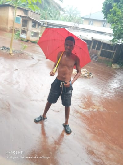 A young Black male stands in the rain holding a red umbrella over his head. He is shirtless and wearing black shorts and black slide sandals. The ground is muddy and wet, with reflections of the sky and surroundings. In the background, there are buildings and trees. The image is geotagged to Umuokwara/Umuezeaga in Akokwa, Nigeria.
