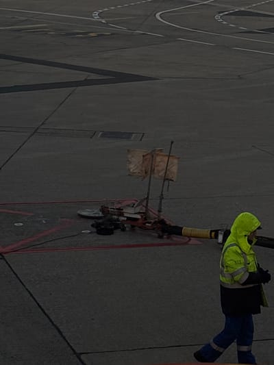 The image depicts an airport ground support operation in Paray-Vieille-Poste, France, likely at Paris-Orly Airport (ORY). The scene is set on a dark gray asphalt tarmac, characterized by various white, yellow, and red painted ground markings for taxiways and operational zones.

In the center-left, a piece of ground support equipment, possibly a fuel hydra-cart, is positioned. This equipment features an orange or red frame, a yellow corrugated hose extending horizontally to the right out of the frame, and two upright poles topped with rectangular, beige fabric warning flags or markers. Attached to the equipment are several black hoses, a silver disc-shaped component, and other mechanical parts. Red lines on the tarmac define a rectangular area around the equipment. Further back on the tarmac, a metal grate or access panel is visible.

In the lower right foreground, a single individual is walking from right to left. This person is wearing high-visibility work attire, including a neon yellow/lime green hooded jacket with reflective gray stripes on the chest and arms, dark blue or black trousers with reflective stripes near the ankles, and dark gloves. The individual's face is partially obscured by the hood, and their gaze is directed towards the left side of the frame. The overall lighting is dim, suggesting either dawn, dusk, or heavily overcast conditions.
