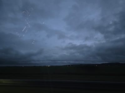 A low-angle shot captures a dark and brooding sky filled with layers of heavy, grey clouds. A thin strip of dark green land stretches across the horizon, punctuated by faint, distant lights. The scene suggests a rural setting viewed through a window, as reflections obscure the upper left corner of the image, indicating movement. Overall, the mood is somber and ominous, reflecting the weather conditions.