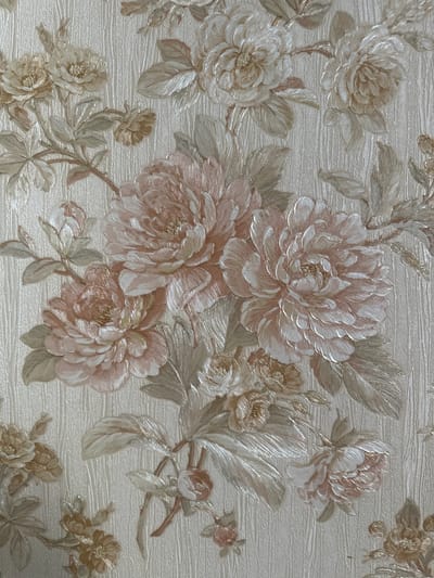 The image displays a close-up of wallpaper featuring a floral pattern. The flowers are large and appear to be peonies or similar blooms, rendered in shades of pink, cream, and gold, with textured detailing on the petals. Accompanying the flowers are various leaves and stems, also textured, in shades of green, beige, and gold. The background of the wallpaper has a subtle vertical ribbed texture. There are no people, actions, or interactions visible in this image, and it does not depict any location in Taraz city administration, Kazakhstan.