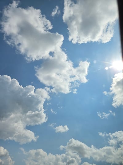 The image is a low-angle shot of a sky in Dalbok Dol, Bulgaria. The sky is blue with scattered cumulus clouds, and the sun is visible as a bright spot at the top of the frame. The clouds are white and fluffy, and they are distributed throughout the frame, occupying the majority of the view. A black object is present in the upper left corner of the frame.