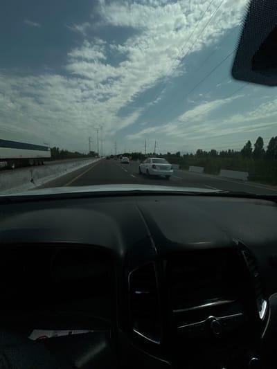 The image captures a view from inside a vehicle looking out at a highway in the city of Istiqlol, Uzbekistan. The sky is cloudy with patches of blue. Multiple lanes of traffic are visible, with several cars and a large truck on the road. Power lines crisscross the sky, supported by tall metal towers. The road is bordered by vegetation and concrete barriers. The interior of the vehicle, including the dashboard and windshield, is also visible.