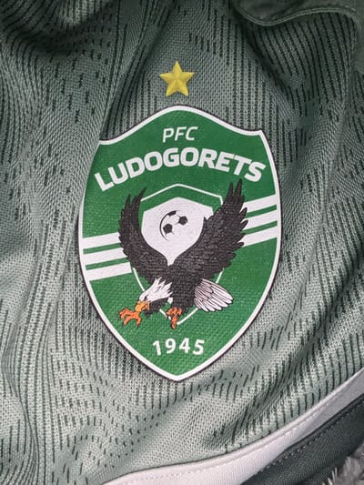 The image depicts a close-up of a green and white PFC Ludogorets soccer jersey. The jersey features the team's emblem, which is a green shield with white accents. The emblem displays the team's name "PFC Ludogorets," a football, and a two-headed eagle. The number "1945" is also present on the badge. A yellow star is to the side of the badge. The jersey is made of a textured material. The location is not evident from the image.