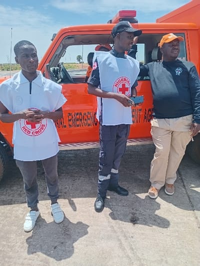 Three men are standing in front of an orange emergency vehicle. The man on the left is wearing a white shirt with a red cross logo and grey pants. The man in the middle is wearing a white vest with a red cross logo, blue pants, and a black cap. The man on the right is wearing a black shirt with a red cross logo, tan pants, and a red cap. They are all standing on concrete. The vehicle is parked in front of a paved road. The location of the image is Katabu, Nigeria, coordinates (latitude: 10.71, longitude: 7.514). The vehicle has the words "National Emergency Management Agency" written on its side. The man on the left is holding his hands clasped in front of him. The man in the middle is holding a black phone in his right hand. The man on the right is standing with his hands on his hips. The background of the image is a blue sky with white clouds.