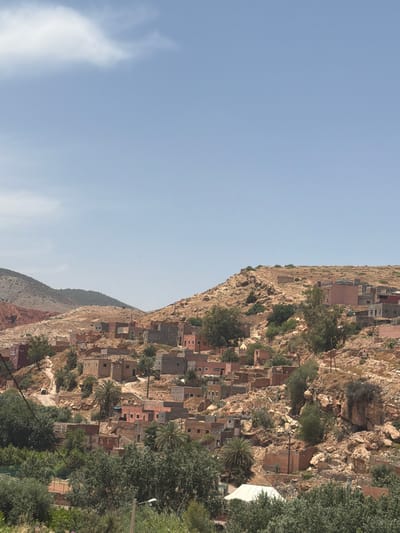 The image captures a vibrant village nestled into the steep, rocky slopes of a mountain under a clear, bright sky. The perspective is a wide shot, looking across at the mountainside settlement.
The sky, occupying the upper left portion of the frame, is a pale, clear blue with a few wispy white clouds, indicating daytime, likely late morning or early afternoon. The dominant feature is the expansive, arid mountain, characterized by its reddish-brown and light tan rock formations and sparse, dry scrub vegetation.
The village buildings are tightly clustered along the contours of the mountain, appearing to be constructed from natural, earthy materials like stone or mud brick, often painted in warm tones of beige, light brown, and soft reddish-pink. These structures are integrated directly into the rugged terrain. Interspersed among the houses are numerous green trees, including distinctive palm trees and other deciduous foliage, providing a striking contrast to the dry mountain and suggesting irrigated areas within the settlement. A notable white tent-like structure is visible among the houses and trees in the mid-right.
Utility poles and lines are visible, indicating modern infrastructure within the traditional setting. Further mountain ranges with similar characteristics can be seen in the background, some appearing hazy. No people are visible, and there is no discernible specific activity taking place; the scene depicts a static landscape. The architecture and landscape elements are consistent with a mountain village in Tahannaout, Morocco. There is no visible text in the image.